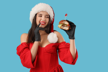 Wall Mural - Pretty young woman in Santa costume with tasty burger on blue background