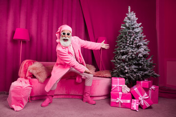 Photo of funny retired man santa claus point tree fur coat boots fluffy nighty hat sunglasses pink christmas party decor interior x-mas