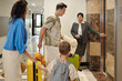 © DragonImages - Family standing near elevator in hotel lobby while employee holding door Family carrying luggage and child wearing backpack, all preparing to enter elevator