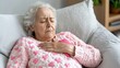 © Iona - An elderly woman with closed eyes rests on a couch, her hand placed on her heart in a moment of introspection.