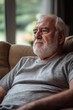 © Vitalii Shkurko - An elderly man with a white beard sits quietly on a couch, lost in thought, enjoying the light filtering in through the windows in a warm living room