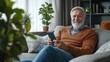© Thuntaree - Happy older man with gray beard casually using smartphone while sitting on cozy sofa surrounded by indoor plants in a bright living room, showing a relaxed lifestyle at home