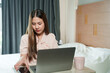 © Ekkasit A Siam - Asian adult woman working at home sitting in cozy bedroom, wearing light pink blouse, holding smartphone and using laptop, warm lighting creating relaxed atmosphere, modern interior design visible.