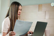 © Ekkasit A Siam - Asian adult woman sitting in cozy bedroom with light wooden walls, while working on laptop, holding paper document, appearing serious, wearing light pink blouse, warm lighting reflecting dedicated