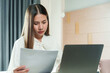 © Ekkasit A Siam - Asian woman holding paper document while working on laptop, white blouse, serious expression, seated in bedroom with modern wooden decor, warm lighting, illustrating work-from-home scenario.