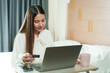 © Ekkasit A Siam - Asian young woman holding credit card, sitting in cozy bedroom, wearing white blouse, smiling while using laptop, representing online shopping and digital payment concept.