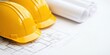 © Kamonwan - Two yellow hard hats on architectural plans and rolled blueprints on a white surface.