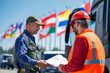 © Iriyalo - Customs officer checking documents with a worker in high-visibility clothing at an international checkpoint with flags in the background