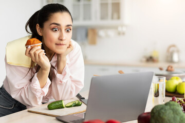 Poster - Exciting Movie Concept. Portrait of terrified Amazed surprised young lady watching film or reality show in kitchen at home, using laptop computer while cooking, eating muffin cake, leaning on counter