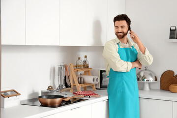 Wall Mural - Handsome bearded man talking by mobile phone near frying pan on stove in kitchen