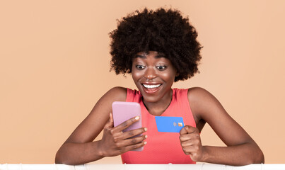 Poster - Joyful Black Woman Shopping Using Smartphone And Credit Card Standing Near Clothing Rack Purchasing Trendy Summer Clothes Over Yellow Background, Studio Shot