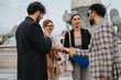 © qunica.com - A group of business people engaged in a lively discussion in an urban setting. They are shaking hands, sharing ideas, and collaborating on strategic solutions in a friendly outdoor environment.