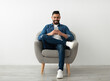 © Prostock-studio - Cheerful young Arab man using smartphone, having online chat, sitting in armchair, browsing social media against white studio wall, blank space. Remote communication, blogging concept