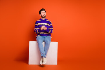Aufkleber - Full body photo of young man sit white cube hold device look empty space dressed striped purple clothes isolated on orange color background