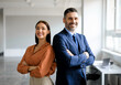 © Prostock-studio - Successful happy businessman and businesswoman standing back to back with arms crossed and smiling at camera, office interior. Partnership concept