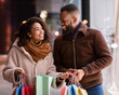 © Prostock-studio - Enjoying Purchases. Portrait of happy smiling black couple holding colorful shopping bags, excited about their new clothes or gift, standing outdoors near mall in the evening, looking at each other