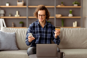 Poster - Happy Guy Using Credit Card And Smartphone Shopping Online In Mobile Application Sitting At Laptop On Sofa At Home. Man Making Payment Via Bank App On Phone Indoors. E-Commerce Concept