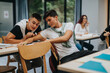 © qunica.com - Two students collaborate at a desk in a modern classroom environment. They share a moment of teamwork and discussion while other students focus on their studies.
