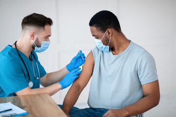 Poster - Medical doctor or nurse giving coronavirus vaccine shot to African American male patient at clinic. Young black man in face mask getting covid-19 vaccine, participating in immunization campaign