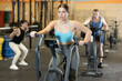 © JackF - Energetic young female athlete training with exercise bike machine near other people gym