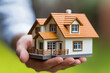 © Pics_With_Love - Hand holding miniature house model symbolizing real estate, property ownership, home insurance, and mortgage concepts, set against a blurred natural green background.