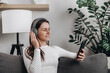 © Yura Yarema - Relaxed happy girl wearing earphones listening to mobile music playing via smartphone app, calm young woman in headphones chilling on cozy sofa enjoying songs podcast holding cell phone at home