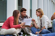 © Gigi Delgado - Three student friends sitting on the ground outside university, studying together after class having a brainstorming using a notebook to write down ideas.