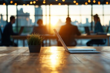 Poster - A laptop placed on a wooden table in an office setting at sunset, with blurred colleagues in the background highlighting a modern, collaborative work environment.