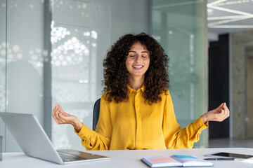 woman meditates at office desk, demonstrating mindfulness with laptop and phone nearby. she is relax