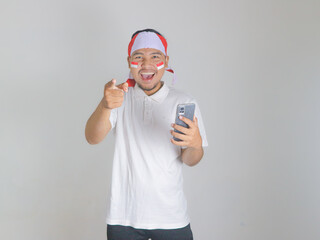 Wall Mural - Excited young Asian men celebrate Indonesian independence day on 17 August while holding mobile phone and pointing forward isolated over white background