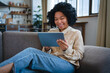 © InsideCreativeHouse - Close up of smiling young African woman relaxing on sofa using tablet computer reading book watching movie media content chatting online talking on video call. Internet communication