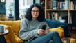 © 99 RIC - A cheerful young Asian woman with glasses relaxes on a colorful couch, holding a smartphone. She's dressed casually in a cream sweater and white pants, surrounded by a cozy home environment with books