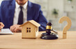 © Studio Romantic - Judge gavel, small wooden toy house and a question mark standing at the desk with a male lawyer working with documents in background. Real estate law, house foreclosure and bankruptcy concept.