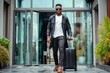 © Zaleman - Black male traveler standing against modern glass building in city, holding suitcase, looking away at camera from below