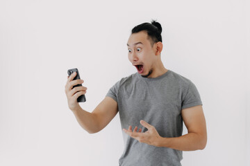 Wall Mural - Shock and wow face Asian man in grey t-shirt use smartphone app on white.