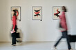 © AnnaStills - Blurred figures passing by modern art pieces on gallery walls showing dynamic forms and vibrant colors creating sense of motion and interaction with the artwork