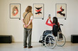 © AnnaStills - Two people in modern gallery observing and photographing abstract art pieces displayed on wall. One person is standing while other is sitting in a wheelchair