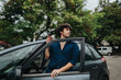 © qunica.com - Young man in a blue shirt exits a parked car surrounded by lush greenery in an open parking space, looking contemplative and relaxed.
