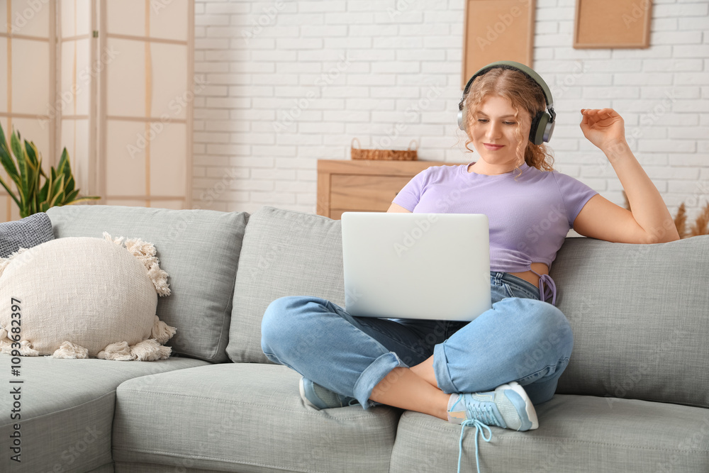 Young woman in headphones using laptop on sofa at home