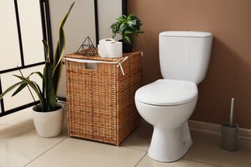 Modern toilet bowl, basket with paper and houseplants near brown wall in restroom