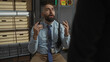 © Krakenimages.com - A bald man with a beard wearing a blue shirt and tie gesticulates while talking, a detective badge visible, in a room with file boxes.