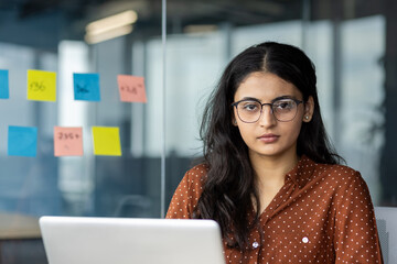 Wall Mural - Confident Latin American businesswoman working on laptop at modern office. She is focused with sticky notes on glass wall behind her. Perfect for business, technology, and leadership themes.