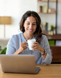 © anandart - Happy mature woman sitting at home office table drinking coffee looking at laptop. Smiling relaxed middle aged older lady hybrid working on computer watching elearning webinar in modern house