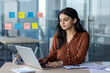 © Liubomir - Latin American woman businesswoman working on laptop in office with glass walls and sticky notes. She wears brown polka dot blouse, demonstrating focus, professionalism, and modern work environment.