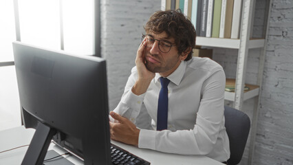 Wall Mural - Man looking tired in office with computer wearing glasses and white shirt beside shelves with books in bright indoor workspace during workday.