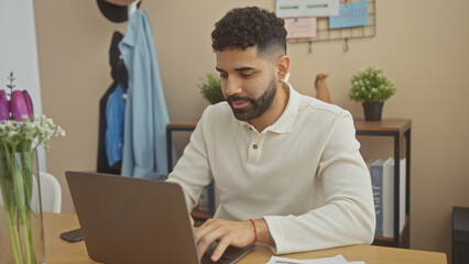 Wall Mural - A focused young hispanic man with a beard working on a laptop at home, conveying a professional yet casual atmosphere.