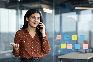 Wall Mural - Latin American woman businesswoman talking on phone in office, smiling and expressing confidence. Engaged in positive communication near glass wall with sticky notes, embodying professional success.