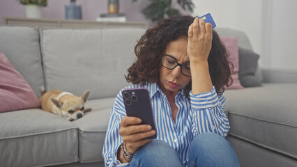 Sticker - Stressed woman holding credit card looks at phone, unaware of chihuahua resting on couch in cozy living room.