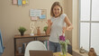 © Krakenimages.com - Happy mature woman dancing alone in a cozy modern home office with a laptop, plants, and decorative items.