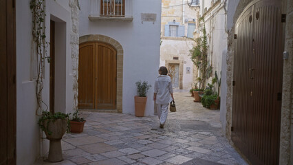 Wall Mural - Young hispanic woman walks through charming narrow streets of polignano a mare, puglia, italy, showcasing beautiful, historic architecture and vibrant, quaint alleyway surroundings.